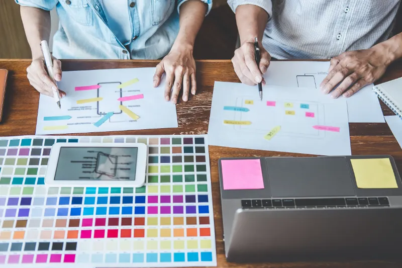Zwei Personen arbeiten an einer Logo Grafik Design Skizze, umgeben von Farbmusterkarten und einem Tablet auf einem Tisch